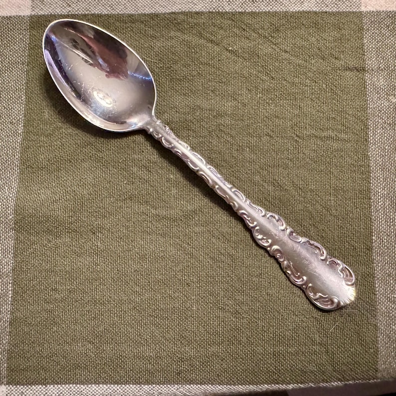 May include: A silver-toned metal spoon featuring a polished oval bowl and a handle decorated with an embossed scrollwork pattern. The handle tapers toward the bowl and widens at the rounded end, showcasing a detailed relief design.