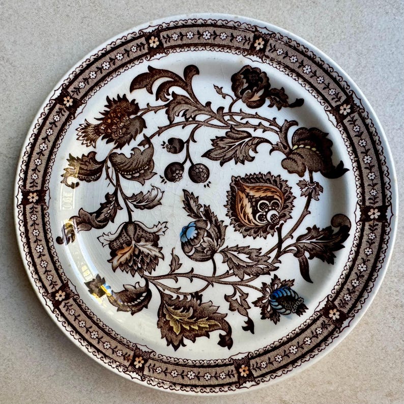 May include: A round ceramic plate with a cream-colored base featuring a dark brown floral and vine pattern. The rim has a repeating geometric border with small white flowers. Two flower buds are accented with light blue and yellow paint.