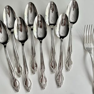 May include: A collection of polished silver spoons and forks arranged on a white surface. The spoons have a rounded bowl and ornate handle design. The forks have four tines. The silverware reflects the surrounding environment.