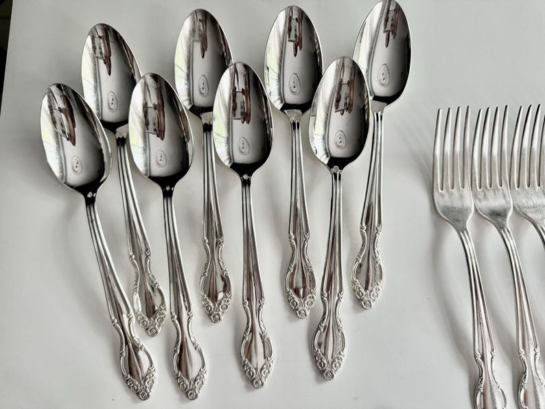 May include: A collection of polished silver spoons and forks arranged on a white surface. The spoons have a rounded bowl and ornate handle design. The forks have four tines. The silverware reflects the surrounding environment.