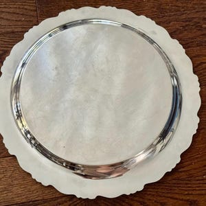 May include: A round silver-toned serving tray with a smooth, reflective center surface and a decorative, scalloped edge. The metal platter features a raised rim and a polished finish that catches the light, creating a bright, metallic appearance.