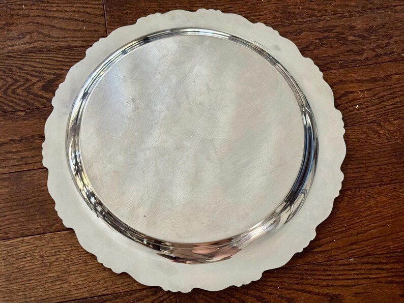 May include: A round silver-toned serving tray with a smooth, reflective center surface and a decorative, scalloped edge. The metal platter features a raised rim and a polished finish that catches the light, creating a bright, metallic appearance.
