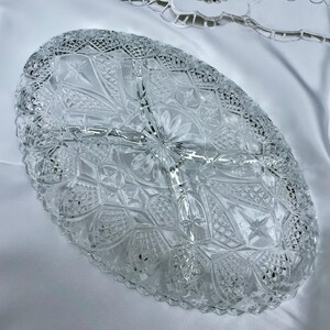 May include: Clear, oval-shaped glass serving dish with a detailed, cut-glass design. The dish is divided into three sections and features a scalloped edge. The intricate pattern includes stars, flowers, and geometric shapes, creating a vintage aesthetic.