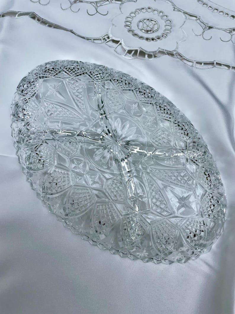 May include: Clear, oval-shaped glass serving dish with a detailed, cut-glass design. The dish is divided into three sections and features a scalloped edge. The intricate pattern includes stars, flowers, and geometric shapes, creating a vintage aesthetic.