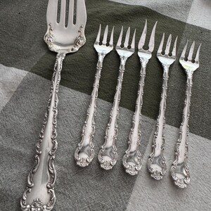 K&ouml;nnte beinhalten: Ein Set aus f&uuml;nf silberfarbenen Metallgabeln mit verzierten, gepr&auml;gten Schn&ouml;rkelmustern an den Griffen. Die Kollektion umfasst eine gro&szlig;e Vorlegegabel mit vier Zinken und vier kleinere Gabeln mit jeweils drei Zinken, alle mit polierter Oberfl&auml;che.