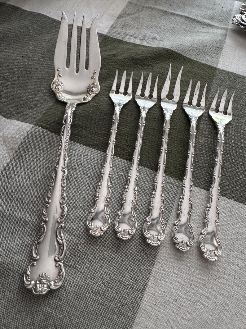 K&ouml;nnte beinhalten: Ein Set aus f&uuml;nf silberfarbenen Metallgabeln mit verzierten, gepr&auml;gten Schn&ouml;rkelmustern an den Griffen. Die Kollektion umfasst eine gro&szlig;e Vorlegegabel mit vier Zinken und vier kleinere Gabeln mit jeweils drei Zinken, alle mit polierter Oberfl&auml;che.