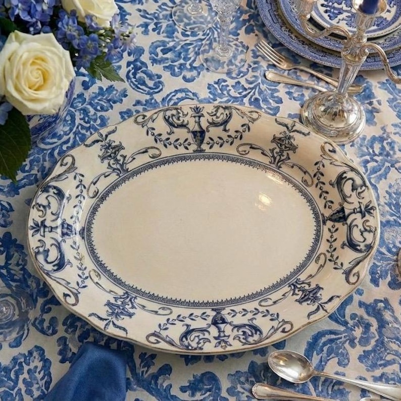 K&ouml;nnte beinhalten: Eine ovale Servierplatte mit wei&szlig;er Mitte und blauem Blumendesign am Rand. Die Platte steht auf einer blau-wei&szlig; gemusterten Tischdecke, mit Blumen und Besteck im Hintergrund.