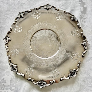 May include: A round silver-toned metal serving tray featuring an ornate scalloped edge with embossed grape cluster motifs. The center and border are decorated with intricate, etched scrollwork and symmetrical urn-shaped patterns.