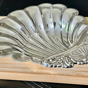 May include: A silver shell-shaped dish with an ornate design on the edge. The dish is sitting on a wooden surface.