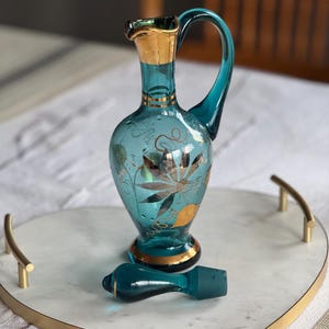May include: A teal blue glass bottle with a gold rim and a gold floral design. The bottle has a stopper and is sitting on a white marble tray with a gold rim.