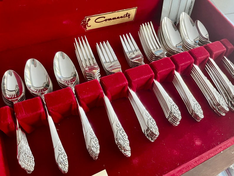 May include: A set of Community silverware, including spoons, forks, and knives, arranged in a red velvet-lined box. The silverware has a floral design on the handles and is displayed neatly. The box has a gold label.