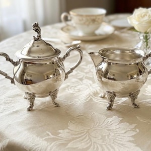 Puede incluir: Un azucarero de plata pulida con tapa a juego y una jarra de leche de plata. Ambas piezas presentan asas curvas ornamentadas y pies de tr&iacute;pode decorativos con detalles intrincados. Las superficies de metal reflejan un acabado brillante.