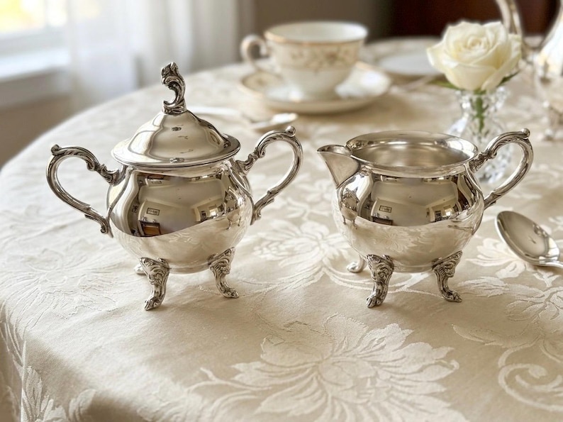 Puede incluir: Un azucarero de plata pulida con tapa a juego y una jarra de leche de plata. Ambas piezas presentan asas curvas ornamentadas y pies de tr&iacute;pode decorativos con detalles intrincados. Las superficies de metal reflejan un acabado brillante.
