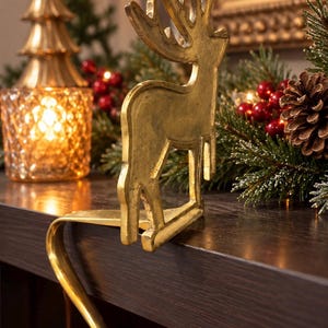 May include: A gold-toned metal stocking holder featuring a standing reindeer silhouette with detailed antlers. The holder has a curved hook design that extends over the edge of a flat surface to support a hanging stocking.