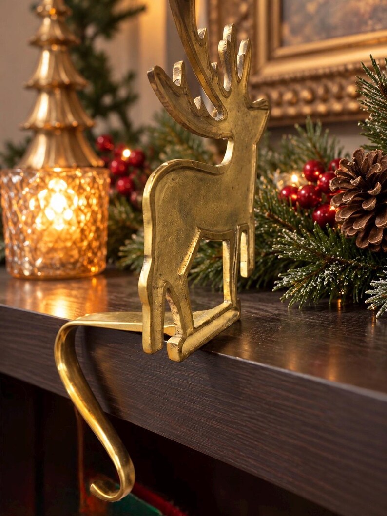 May include: A gold-toned metal stocking holder featuring a standing reindeer silhouette with detailed antlers. The holder has a curved hook design that extends over the edge of a flat surface to support a hanging stocking.