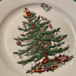 以下が含まれることがあります： 緑色の縁取りのある白いセラミック製のプレートには、手描きのクリスマスツリーが描かれており、オーナメント、プレゼント、そして頂上には小さな人形が飾られています。ツリーは緑の葉のガーランドで囲まれています。
