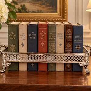 May include: A rectangular silver-toned metal tray with an ornate lattice-patterned side and decorative feet, holding eight hardcover books standing upright. The book spines feature titles including The Garden Book, At Home With Plants, The Kinfolk Home, A History of Art, The Natural World, French Country Living, Poems, and Travels and Discoveries.