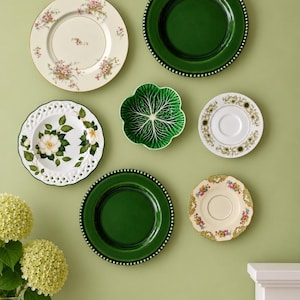 May include: A collection of six decorative plates and a bowl arranged on a wall. The set includes two solid dark green plates with beaded edges, a white plate with pink floral patterns, a white plate with a green floral border, a white plate with a rose illustration and lattice edge, a small floral saucer, and a green cabbage-shaped bowl.
