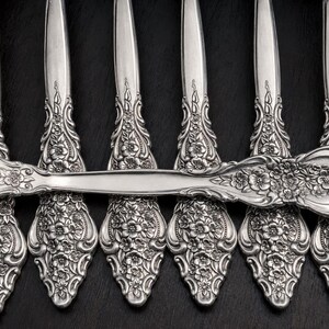 May include: A set of silver-toned metal spoons featuring an intricate embossed floral pattern on the handles. The design includes detailed flowers and scrolling leaves that extend from the base of the handle up to the neck of each spoon.