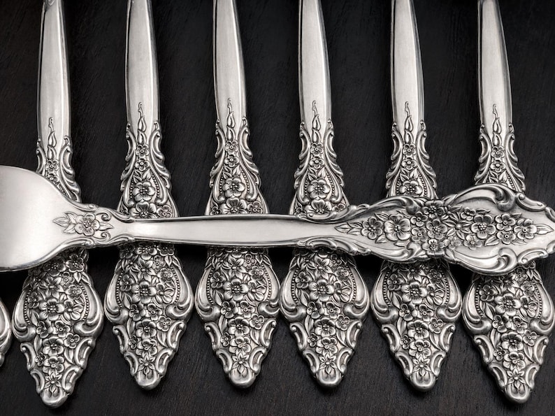 May include: A set of silver-toned metal spoons featuring an intricate embossed floral pattern on the handles. The design includes detailed flowers and scrolling leaves that extend from the base of the handle up to the neck of each spoon.