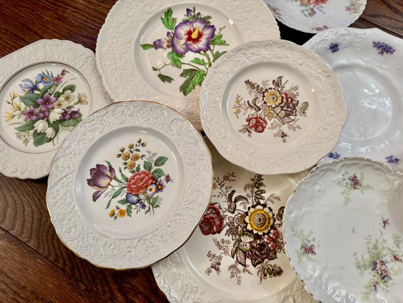 May include: A collection of seven ceramic plates featuring various floral patterns, including depictions of purple hibiscus, mixed wildflowers, and roses. Each plate has a white base with embossed decorative rims and scalloped edges.