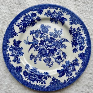 May include: A round ceramic plate featuring a white base with a detailed blue transferware pattern. The design includes central floral arrangements and two birds, surrounded by a wide border of intricate floral motifs and geometric lattice.
