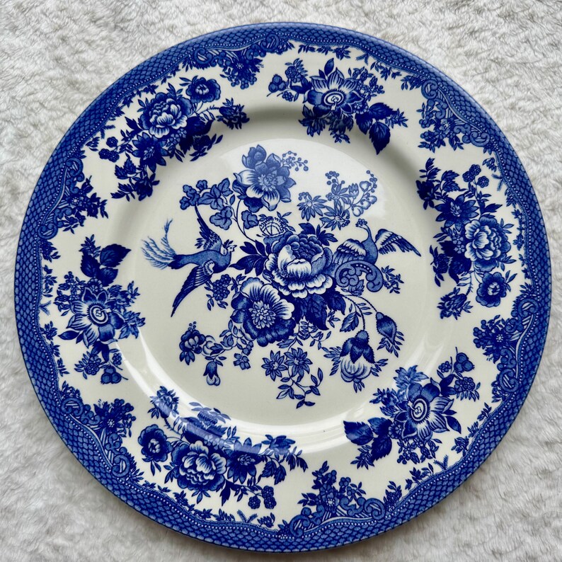 May include: A round ceramic plate featuring a white base with a detailed blue transferware pattern. The design includes central floral arrangements and two birds, surrounded by a wide border of intricate floral motifs and geometric lattice.