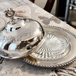 May include: A silver-plated butter dish with a domed lid and a glass insert. The lid features ornate floral engravings and a decorative handle. The dish sits on a silver-plated plate with a beaded edge. The butter dish is open, revealing the glass insert.