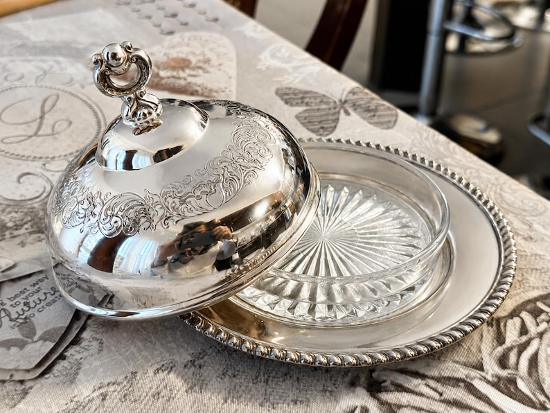 May include: A silver-plated butter dish with a domed lid and a glass insert. The lid features ornate floral engravings and a decorative handle. The dish sits on a silver-plated plate with a beaded edge. The butter dish is open, revealing the glass insert.