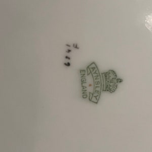 May include: Close-up of a white porcelain plate with the green Aynsley England logo and crown. The plate has the handwritten numbers "6749" and the letter "F" in black ink.