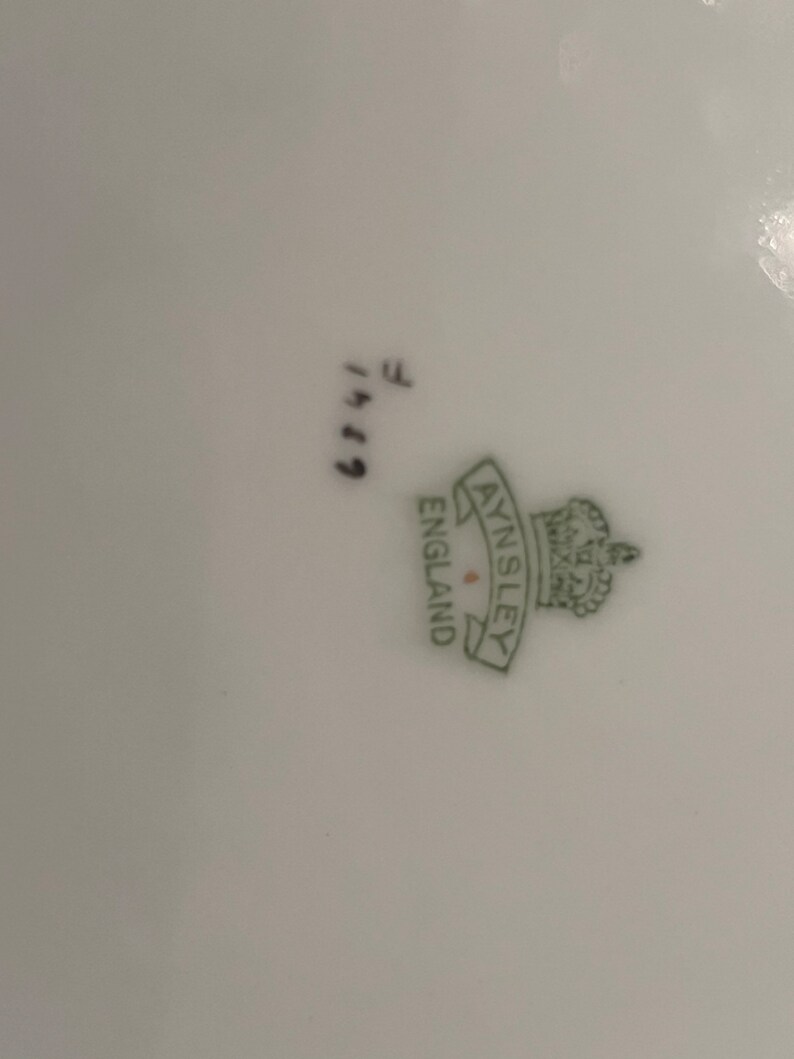 May include: Close-up of a white porcelain plate with the green Aynsley England logo and crown. The plate has the handwritten numbers "6749" and the letter "F" in black ink.