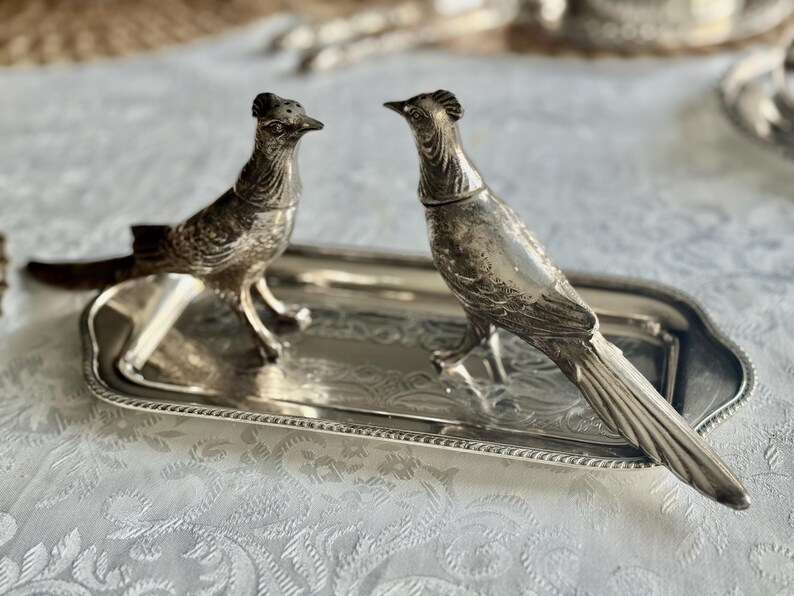 May include: A pair of silver-colored bird-shaped salt and pepper shakers on a matching rectangular tray. The birds are detailed with textured feathers and stand facing each other. The tray has a decorative border.