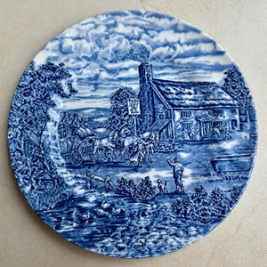 May include: A round ceramic plate featuring a blue and white transferware pattern. The scene depicts a horse-drawn carriage arriving at a thatched-roof inn labeled The Red Lion, with a person and a dog standing in the foreground near a stream.