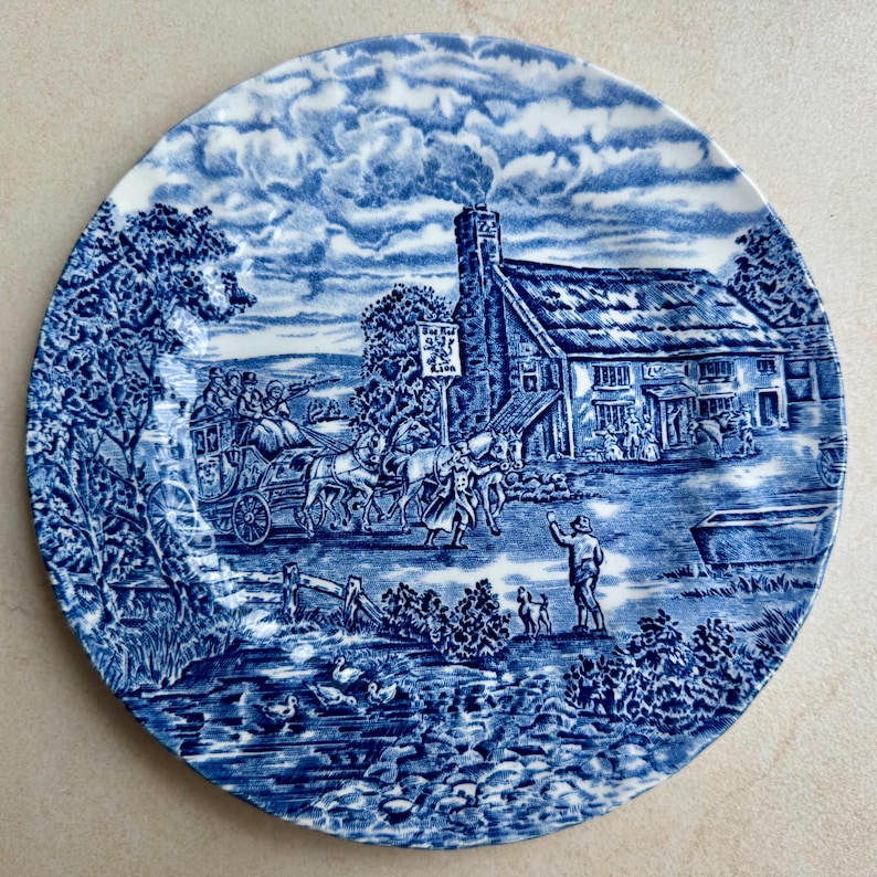 May include: A round ceramic plate featuring a blue and white transferware pattern. The scene depicts a horse-drawn carriage arriving at a thatched-roof inn labeled The Red Lion, with a person and a dog standing in the foreground near a stream.