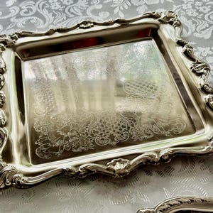 Puede incluir: Una bandeja cuadrada de metal plateado con un borde elevado y ornamentado que presenta intrincados adornos de volutas y detalles en forma de concha. El centro tiene una superficie pulida y reflectante con un sutil patr&oacute;n grabado.