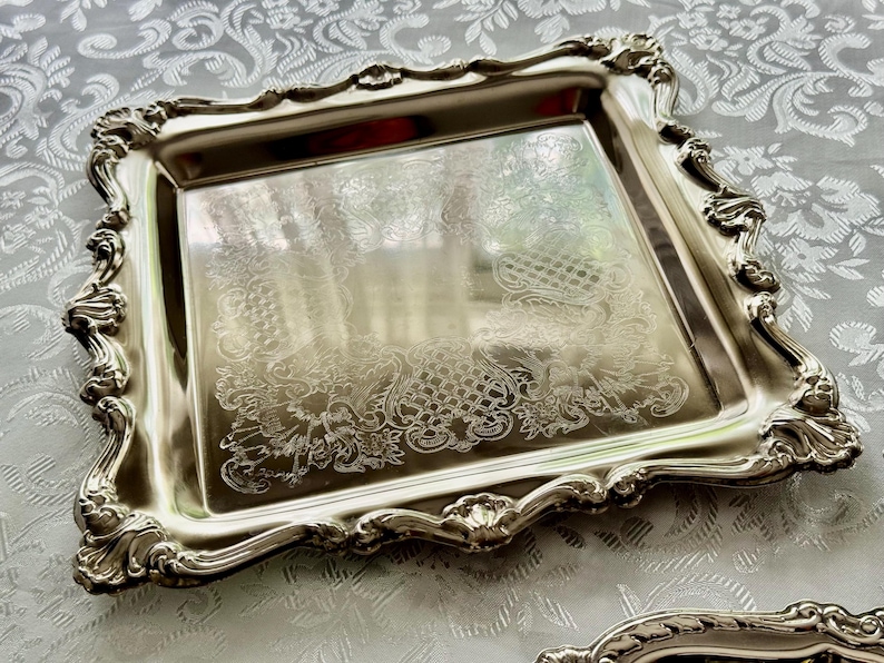 Puede incluir: Una bandeja cuadrada de metal plateado con un borde elevado y ornamentado que presenta intrincados adornos de volutas y detalles en forma de concha. El centro tiene una superficie pulida y reflectante con un sutil patr&oacute;n grabado.
