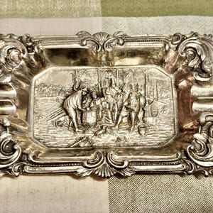 May include: A rectangular silver-toned metal tray featuring an ornate, scrolled border with shell-like motifs. The center depicts a detailed relief scene of several figures gathered around a barrel, with various objects scattered on the ground.