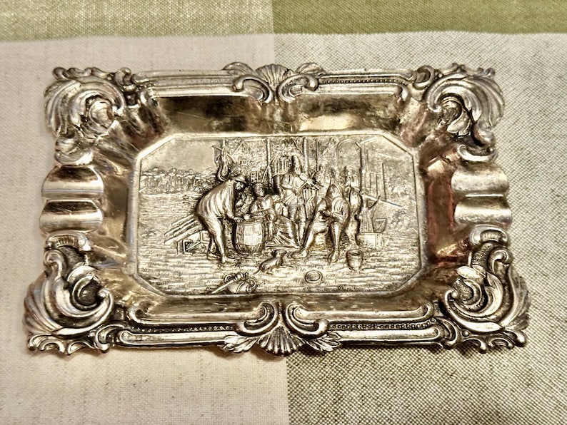 May include: A rectangular silver-toned metal tray featuring an ornate, scrolled border with shell-like motifs. The center depicts a detailed relief scene of several figures gathered around a barrel, with various objects scattered on the ground.
