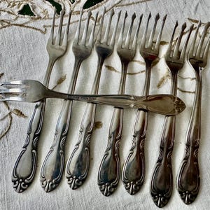May include: A collection of antique silver forks with ornate handles, arranged on a white tablecloth with a floral pattern. The forks are of varying sizes and designs, showcasing a vintage aesthetic.