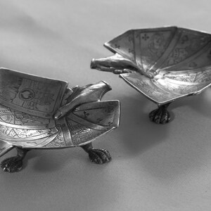 K&ouml;nnte beinhalten: Zwei antike, silberfarbene Aschenbecher in Form einer Schildkr&ouml;te mit aufwendigen Gravuren auf dem Panzer und Krallenf&uuml;&szlig;en. Die Aschenbecher sind auf einer neutralen Oberfl&auml;che platziert. Die Aschenbecher sind aus Metall.