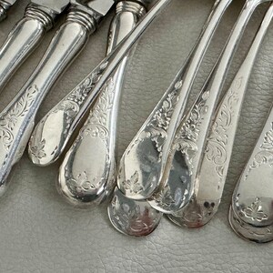 May include: Close-up of antique silverware, including spoons and knives. The silver utensils have ornate floral and leaf patterns etched into the handles and bowls. The silverware is arranged on a light gray surface, reflecting the light.