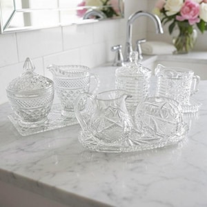 May include: A collection of clear pressed glass tableware including two lidded sugar bowls, two cream pitchers, and two small serving trays. Each piece features intricate geometric diamond and star patterns with a reflective, transparent finish.