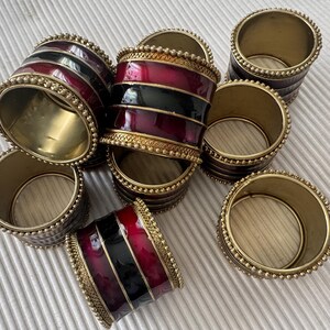 May include: A selection of brass napkin rings. Some rings feature a red and black enamel design with gold beaded trim. Other rings are plain brass with a beaded edge. The napkin rings are arranged on a white surface.