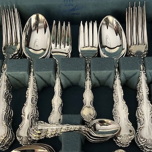 May include: A set of ornate silver-colored flatware, including forks, spoons, and knives, arranged in a fitted case. The handles feature a detailed, scrolling design. The silverware is neatly organized, suggesting a formal dining setting.