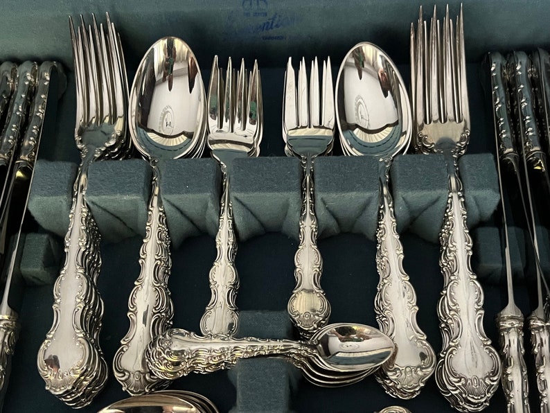 May include: A set of ornate silver-colored flatware, including forks, spoons, and knives, arranged in a fitted case. The handles feature a detailed, scrolling design. The silverware is neatly organized, suggesting a formal dining setting.