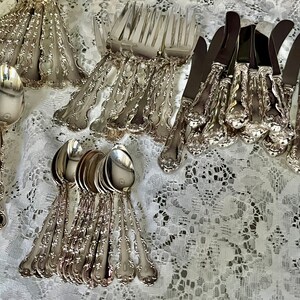 May include: A collection of silver-toned flatware including forks, spoons, and knives with ornate, embossed handles. The utensils feature polished metal surfaces and intricate decorative detailing on the stems, arranged in groups on a white lace surface.
