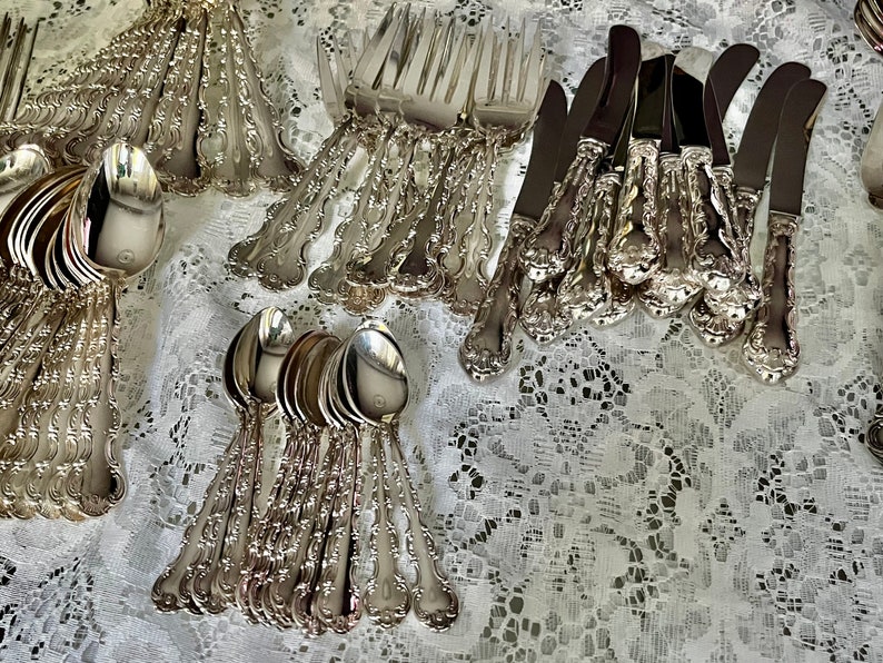 May include: A collection of silver-toned flatware including forks, spoons, and knives with ornate, embossed handles. The utensils feature polished metal surfaces and intricate decorative detailing on the stems, arranged in groups on a white lace surface.