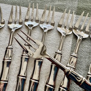 May include: A collection of silver forks with a bamboo-style handle design. The forks are arranged on a light-colored, textured surface. The tines of the forks are visible, and the handles have a detailed, textured appearance.