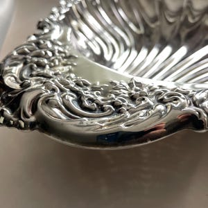 K&ouml;nnte beinhalten: Eine muschelf&ouml;rmige Schale aus silberfarbenem Metall. Die Schale verf&uuml;gt &uuml;ber ein tiefes, geriffeltes Innendesign und einen breiten, verzierten Rand, der mit komplexen, erhabenen Schn&ouml;rkeln und Blumenmustern entlang der geschwungenen Au&szlig;enkante versehen ist.
