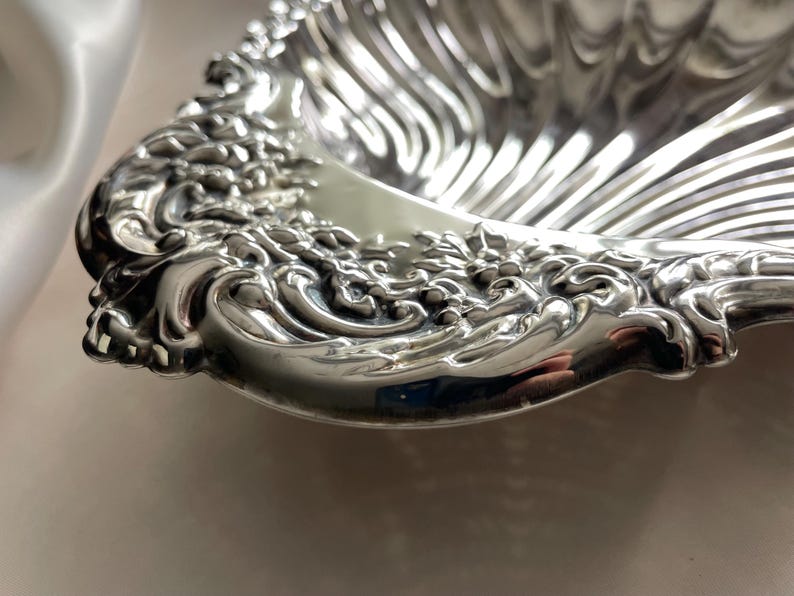 K&ouml;nnte beinhalten: Eine muschelf&ouml;rmige Schale aus silberfarbenem Metall. Die Schale verf&uuml;gt &uuml;ber ein tiefes, geriffeltes Innendesign und einen breiten, verzierten Rand, der mit komplexen, erhabenen Schn&ouml;rkeln und Blumenmustern entlang der geschwungenen Au&szlig;enkante versehen ist.